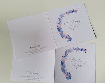 Könnte beinhalten: Eine leere Grußkarte mit einem Aquarellkranz aus blauen und rosa Blumen. Der Text "Thinking of you" ist in Kursivschrift auf der Vorderseite der Karte geschrieben.