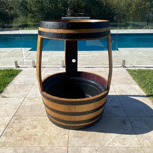 K&ouml;nnte beinhalten: Ein Holzfass-Pflanzgef&auml;&szlig; mit schwarzen Metallb&auml;ndern und einem schwarzen Plastikfutter. Der Pflanzer steht auf einer Terrasse mit einem Schwimmbad im Hintergrund.