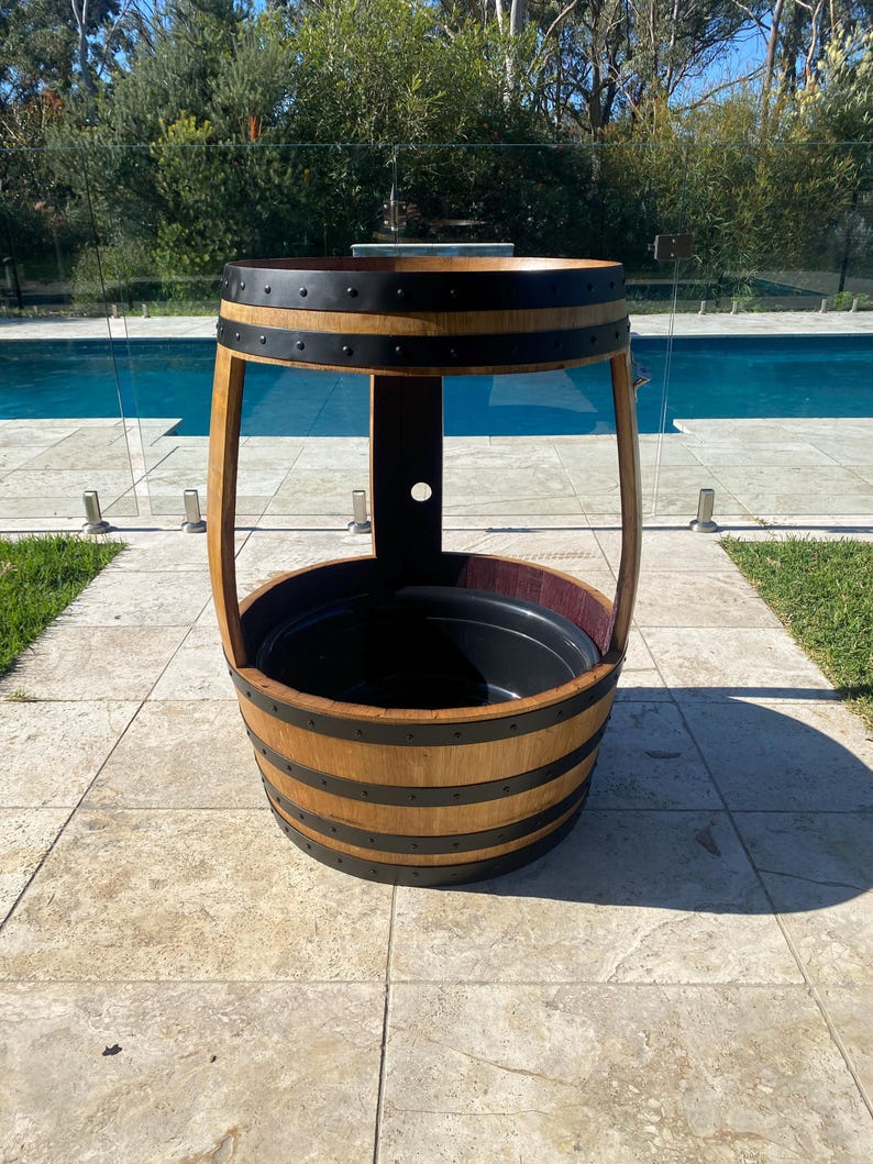 K&ouml;nnte beinhalten: Ein Holzfass-Pflanzgef&auml;&szlig; mit schwarzen Metallb&auml;ndern und einem schwarzen Plastikfutter. Der Pflanzer steht auf einer Terrasse mit einem Schwimmbad im Hintergrund.