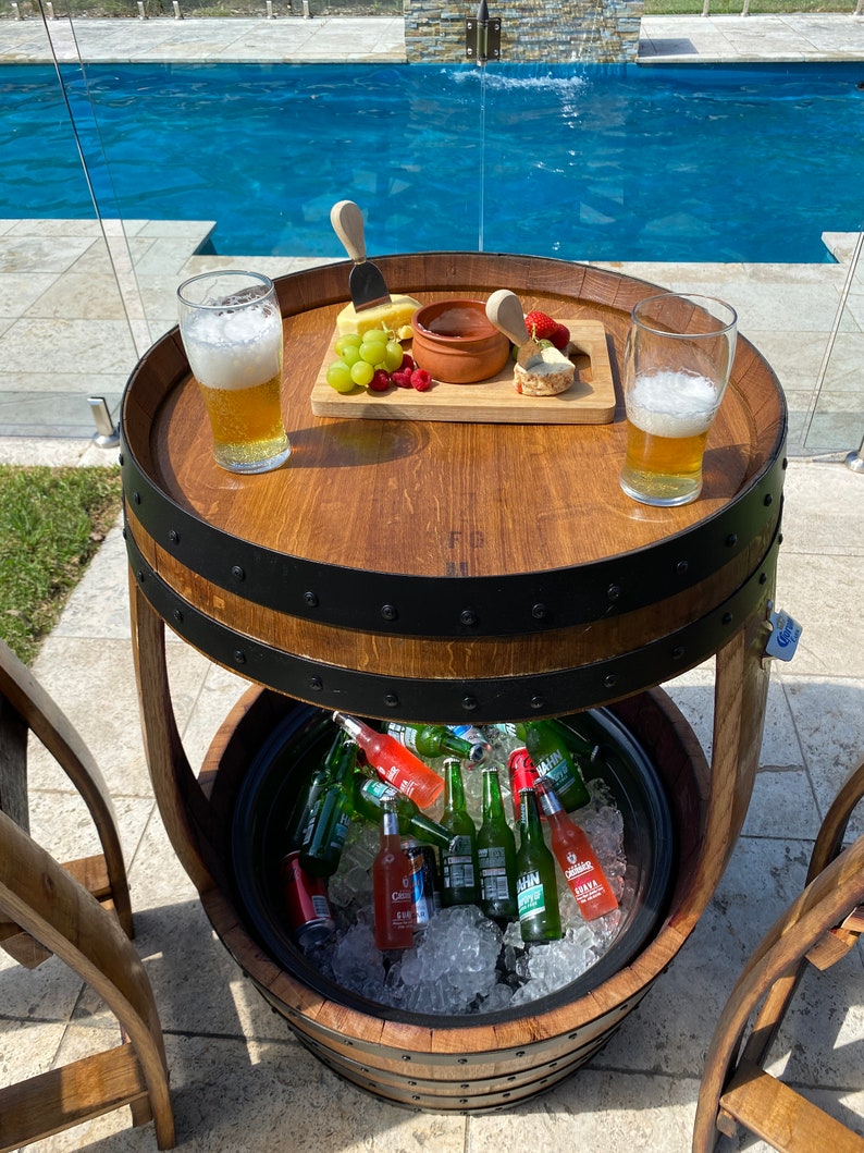 K&ouml;nnte beinhalten: Ein Holzfass-Tisch mit integriertem K&uuml;hlfach. Der Tisch hat eine obere Ablage mit einem Aufschnittbrett, zwei Gl&auml;sern Bier und einer kleinen Sch&uuml;ssel Dip. Das K&uuml;hlfach ist mit Eis und verschiedenen Flaschen Bier und Limonade gef&uuml;llt.