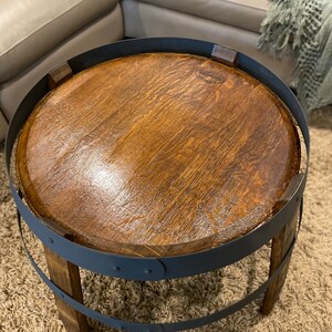 K&ouml;nnte beinhalten: Ein runder Holztisch mit einem schwarzen Metallrahmen. Die Tischplatte ist aus recyceltem Holz gefertigt und hat eine dunkelbraune Farbe. Der Metallrahmen hat zwei B&auml;nder um die Tischplatte und zwei B&auml;nder um die Basis. Der Tisch steht auf einem hellbraunen Teppich.