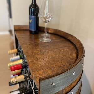 May include: A wooden wine rack with a half-barrel top, featuring a bottle of red wine and two wine glasses. The rack is made of dark wood with metal bands and has a wire grid for storing bottles.
