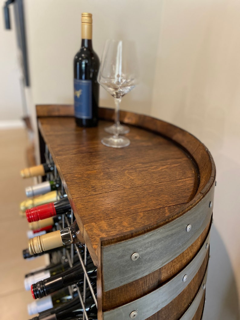 May include: A wooden wine rack with a half-barrel top, featuring a bottle of red wine and two wine glasses. The rack is made of dark wood with metal bands and has a wire grid for storing bottles.