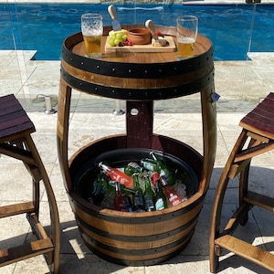 K&ouml;nnte beinhalten: Ein Holzfass-Tisch mit integriertem K&uuml;hlfach. Der Tisch hat eine schwarze Tischplatte mit einem Holzrand und einem schwarzen Metallband. Das K&uuml;hlfach ist mit Eis und Flaschen Bier und Limonade gef&uuml;llt. Der Tisch steht neben einem Schwimmbad.