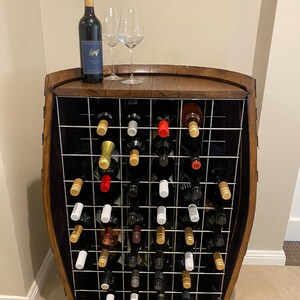 May include: A wooden wine rack with a metal grid holds multiple bottles of wine. The rack is shaped like a barrel and has a wooden top. There are two wine glasses on the top of the rack.