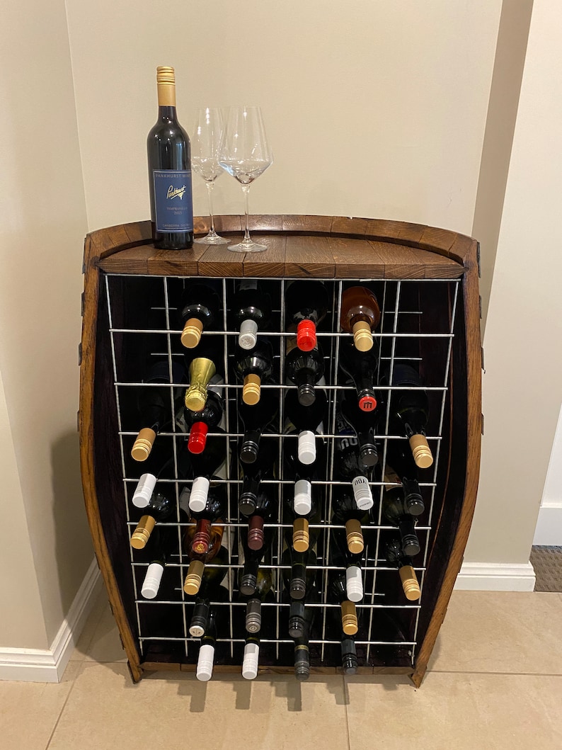 May include: A wooden wine rack with a metal grid holds multiple bottles of wine. The rack is shaped like a barrel and has a wooden top. There are two wine glasses on the top of the rack.