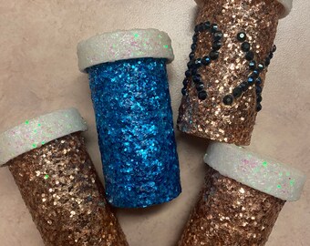 May include: Four small, round, glitter-covered containers with white lids. The containers are filled with different colours of glitter, including gold, blue, and a sparkly white.