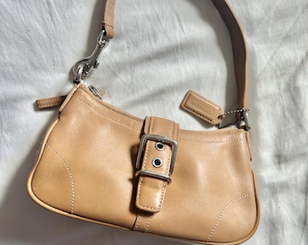 May include: A tan leather satchel handbag with a silver buckle and a silver strap. The bag has a small silver tag with the word "Coach" on it.