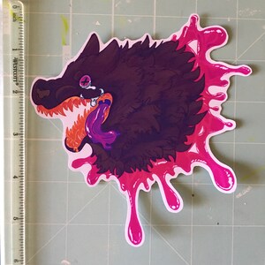 May include: A sticker of a black wolf with a purple tongue sticking out, dripping with pink paint. The wolf has a purple eye and a white tooth.