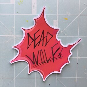 May include: A red and white sticker with a white outline in the shape of a bursting star. The sticker says "DEAD WOLF" in black marker.