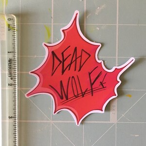 May include: Red and white sticker with a black outline in the shape of a splattered paint splatter. The text "DEAD WOLF" is written in black inside the splatter.