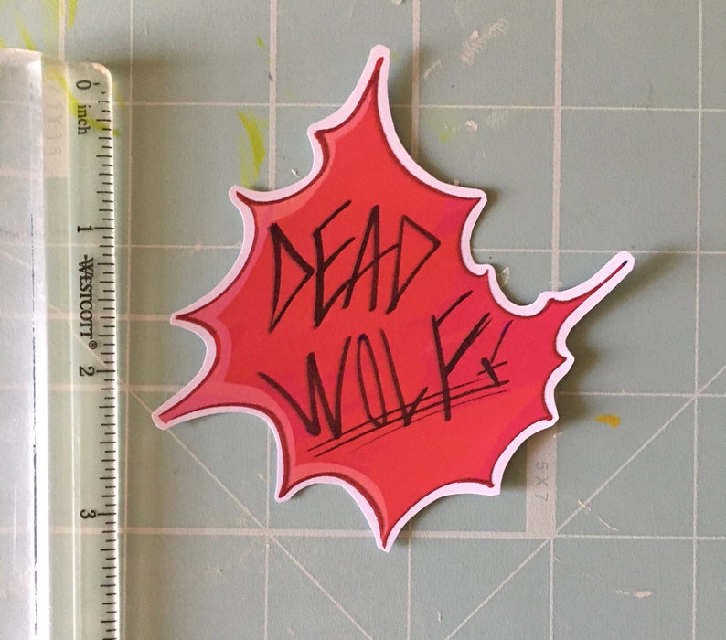 May include: Red and white sticker with a black outline in the shape of a splattered paint splatter. The text "DEAD WOLF" is written in black inside the splatter.