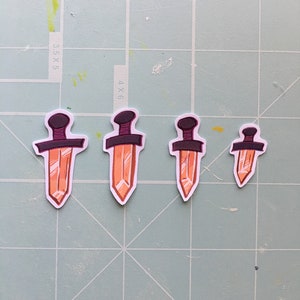 May include: Four orange crystal dagger stickers with black handles. The stickers are arranged in a row from largest to smallest.