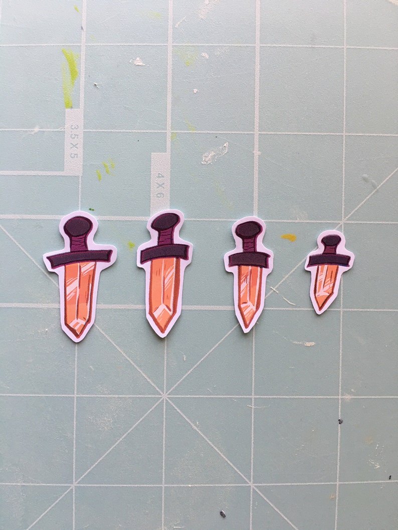 May include: Four orange crystal dagger stickers with black handles. The stickers are arranged in a row from largest to smallest.