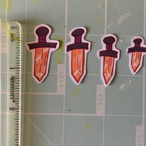 May include: Four orange crystal dagger stickers of varying sizes. The stickers are on a green cutting mat with a ruler in inches.
