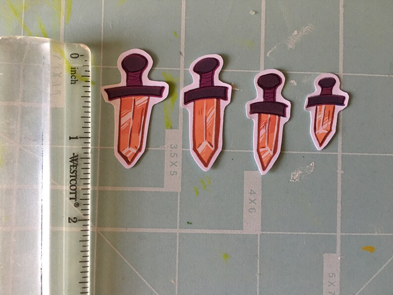 May include: Four orange crystal dagger stickers of varying sizes. The stickers are on a green cutting mat with a ruler in inches.