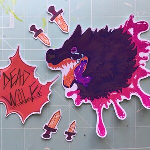 May include: A set of five stickers featuring a black wolf with a pink splatter design, three orange and brown daggers, and a red and white sticker with the text "DEAD WOLF".