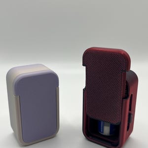 May include: Two 3D printed cases, one is purple and the other is red. The red case is open and has a small white object inside.