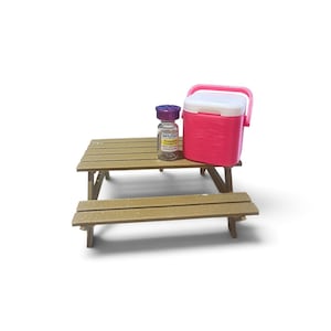 May include: A miniature brown picnic table with a small glass vial featuring a purple cap and a yellow label, alongside a bright pink and white portable cooler. The vial label displays the text USIA PHARMACY and Tirzepatide 12.5mg/0.5ml.