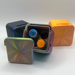 May include: Three small, square, colorful containers with a geometric pattern. One container is open, revealing four small vials inside. The vials are clear glass with blue and orange liquid.