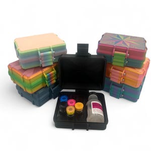 May include: A collection of rectangular storage cases in various colors and patterns, including solid pastel, rainbow gradient, and geometric designs. One open black case in the foreground contains five small colored vials and a bottle of bacteriostatic water.