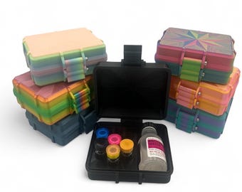 May include: A collection of rectangular storage cases in various colors and patterns, including solid pastel, rainbow gradient, and geometric designs. One open black case in the foreground contains five small colored vials and a bottle of bacteriostatic water.