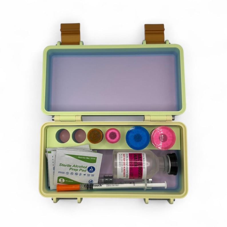 May include: Open, light green medical kit with a purple interior. The kit contains various medical supplies, including vials, syringes, alcohol prep pads, and colored containers. The kit has brown clasps and hinges.