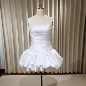 May include: A white strapless dress with a fitted bodice and a ruffled skirt. The dress is displayed on a mannequin against a neutral background. The dress appears to be made of a satin-like material.