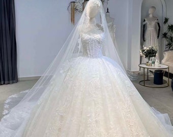 May include: A white ball gown wedding dress featuring a sweetheart neckline, a corset-style bodice with intricate lace embroidery, and a voluminous floor-length skirt with a long train. A sheer, floor-length veil covers the head and shoulders.