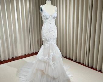 May include: A white wedding dress with a fitted bodice and a flowing skirt. The bodice is covered in delicate floral embroidery and the skirt has multiple layers of tulle.