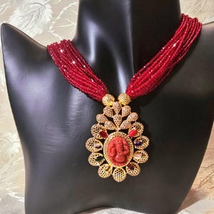 Può includere: Una collana di perline rosse con un ciondolo dorato che presenta un design dettagliato e una pietra preziosa rossa. Il ciondolo ha un'immagine in rilievo di una divinità con pietre preziose colorate.