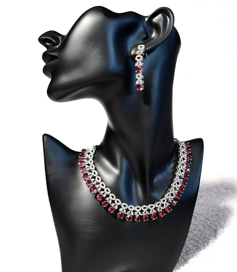 Puede incluir: Un collar y pendientes de plata con una fila de piedras rojas y transparentes. El collar est&aacute; en un busto de maniqu&iacute; negro.