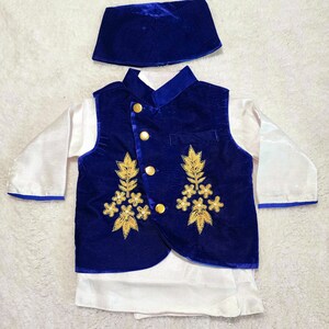 K&ouml;nnte beinhalten: Ein dreiteiliges Kinderoutfit, bestehend aus einer k&ouml;nigsblauen Samtweste mit goldener Stickerei und Kn&ouml;pfen, einer passenden M&uuml;tze und einem wei&szlig;en Langarmhemd. Die Weste hat einen Stehkragen und wird mit einer wei&szlig;en Hose kombiniert.
