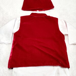 K&ouml;nnte beinhalten: Ein rotes Samt-Westen- und passendes M&uuml;tzen-Set &uuml;ber einem wei&szlig;en Langarmshirt. Die Weste hat ein klassisches Design, und die M&uuml;tze hat eine einfache, abgerundete Form. Das Outfit ist wahrscheinlich f&uuml;r ein Kind.