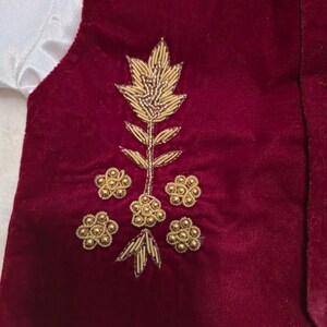 K&ouml;nnte beinhalten: Eine weinrote Samtweste mit goldener Blumenstickerei. Die Stickerei zeigt einen zentralen Stiel mit Bl&auml;ttern und mehrere Blumengruppen. Ein wei&szlig;er Satin&auml;rmel ist auf der linken Seite sichtbar.