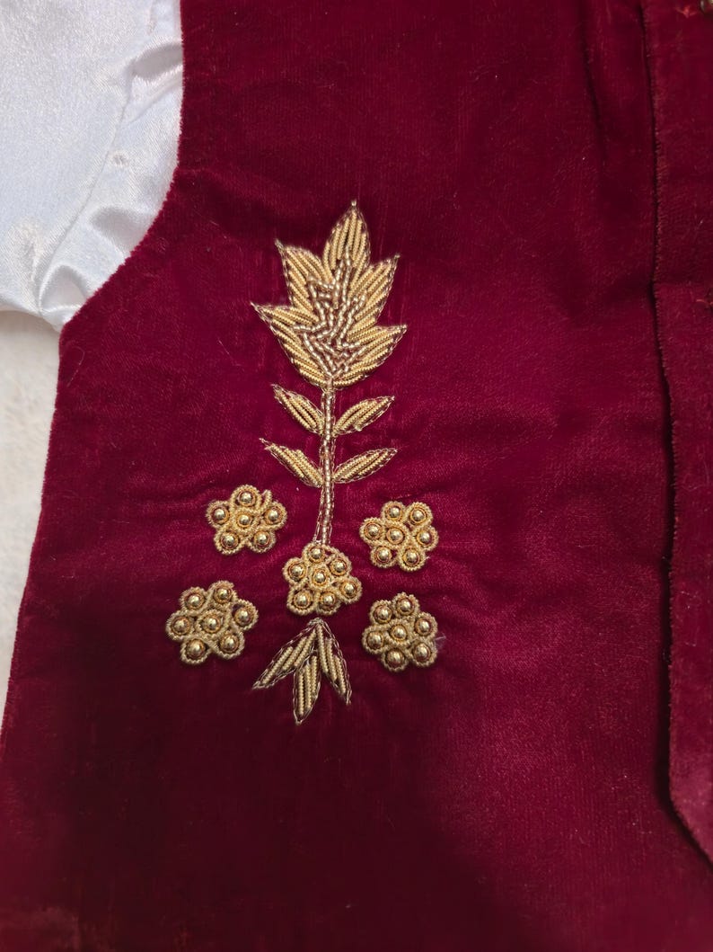 K&ouml;nnte beinhalten: Eine weinrote Samtweste mit goldener Blumenstickerei. Die Stickerei zeigt einen zentralen Stiel mit Bl&auml;ttern und mehrere Blumengruppen. Ein wei&szlig;er Satin&auml;rmel ist auf der linken Seite sichtbar.