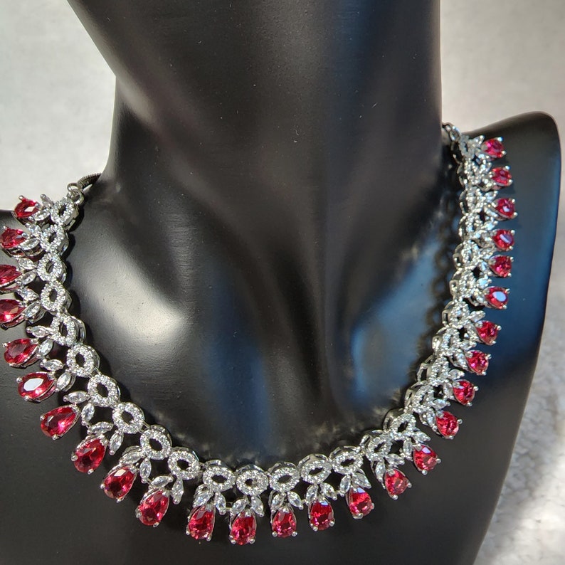 Puede incluir: Un collar de plata con un dise&ntilde;o delicado que presenta peque&ntilde;as piedras transparentes y piedras rojas m&aacute;s grandes en forma de l&aacute;grima.