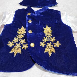 K&ouml;nnte beinhalten: Ein formelles Outfit f&uuml;r ein Kind, bestehend aus einer k&ouml;nigsblauen Samtweste mit goldener Stickerei und goldenen Kn&ouml;pfen. Das Outfit beinhaltet eine passende Kappe und ein wei&szlig;es Hemd mit langen &Auml;rmeln. Die Weste hat ein florales Design.