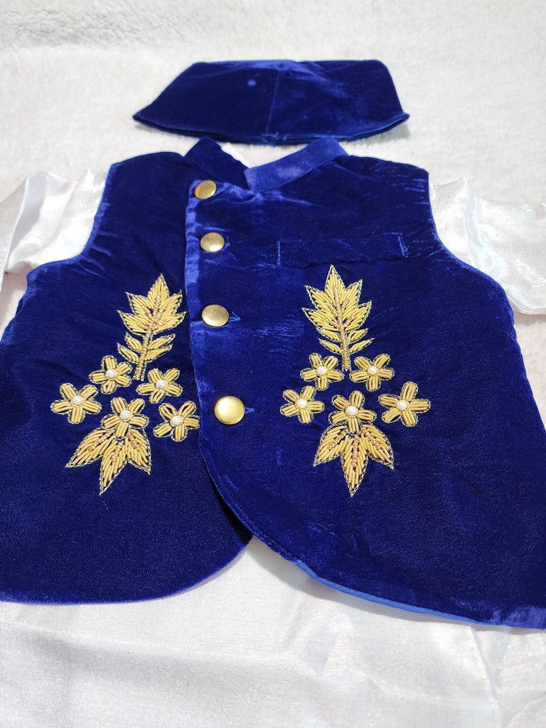 K&ouml;nnte beinhalten: Ein formelles Outfit f&uuml;r ein Kind, bestehend aus einer k&ouml;nigsblauen Samtweste mit goldener Stickerei und goldenen Kn&ouml;pfen. Das Outfit beinhaltet eine passende Kappe und ein wei&szlig;es Hemd mit langen &Auml;rmeln. Die Weste hat ein florales Design.