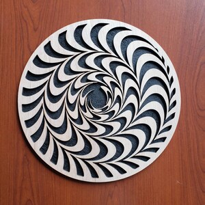 May include: A circular wooden wall decoration featuring a black and white swirling pattern. The pattern is made up of a series of curved lines that create a hypnotic effect.
