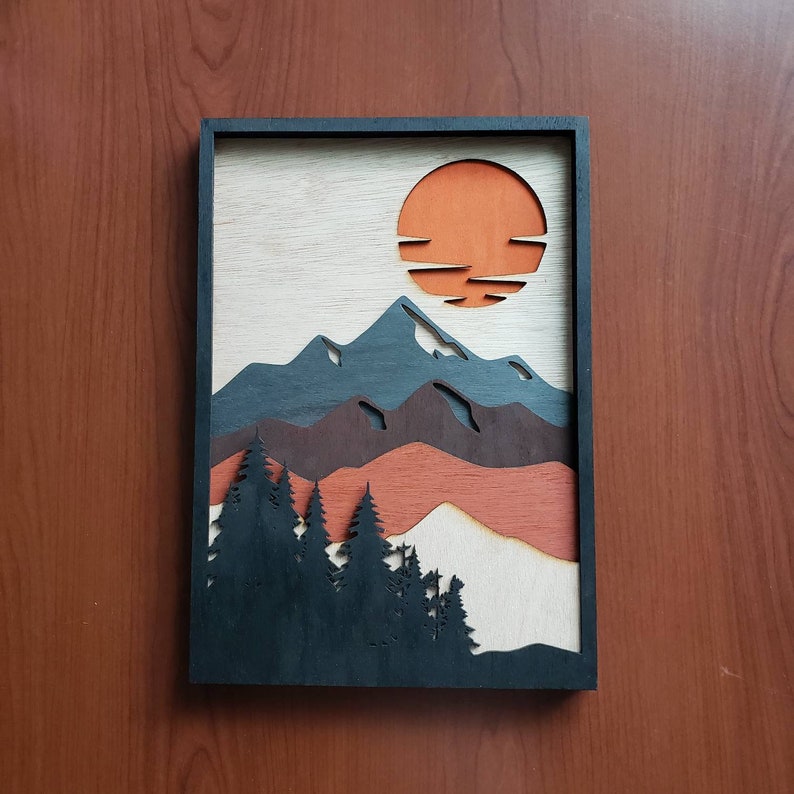 May include: A framed wood wall art piece with a layered design of mountains, trees, and a sunset. The mountains are blue, brown, and red, and the trees are black. The sunset is orange and has a layered design of horizontal lines.