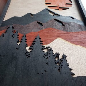 May include: A wood wall art piece with a layered design of mountains, a sunset, and a forest of trees. The mountains are black, the sunset is red and orange, and the trees are black.