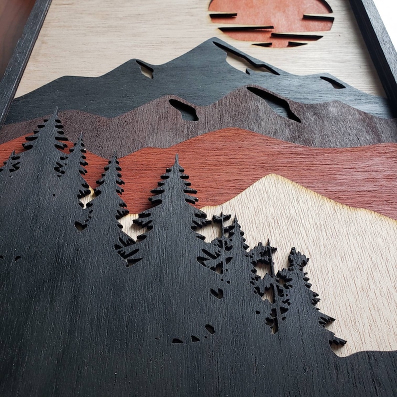 May include: A wood wall art piece with a layered design of mountains, a sunset, and a forest of trees. The mountains are black, the sunset is red and orange, and the trees are black.