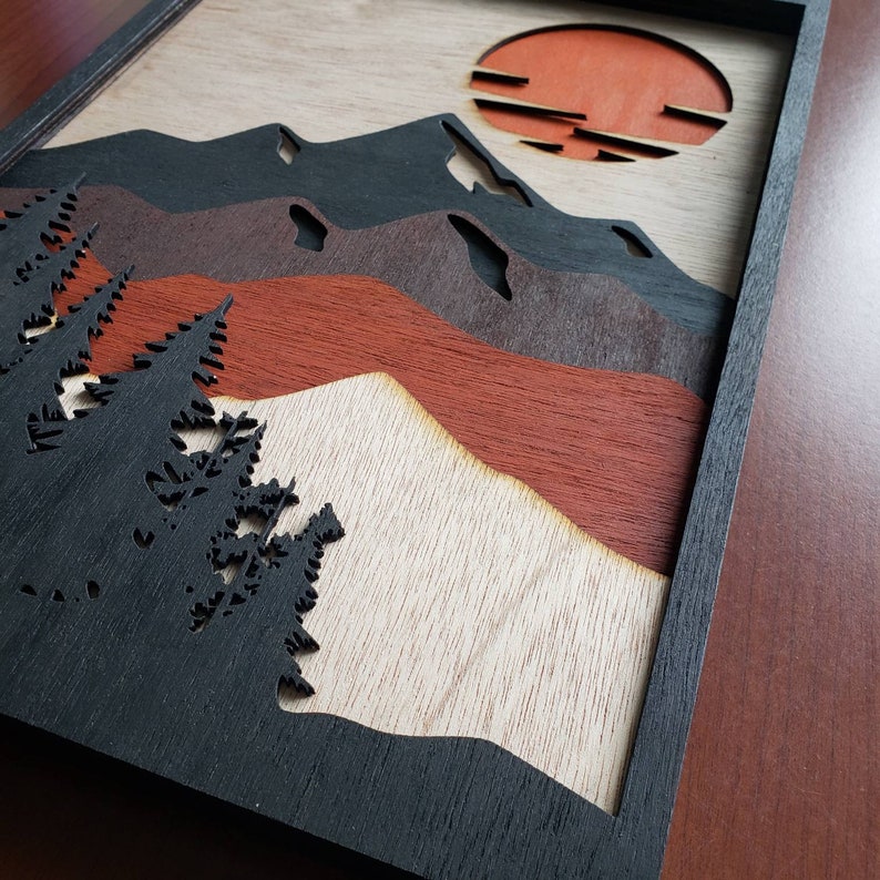 May include: A wooden wall art piece with a black frame. The art features a layered design of mountains, a sunset, and trees. The mountains are in shades of brown and red, the sunset is orange, and the trees are black.