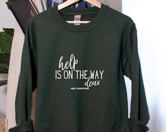 May include: A dark green sweatshirt with the text "help is on the way dear" in white lettering. The text is in a cursive font. The sweatshirt is hanging on a hanger.