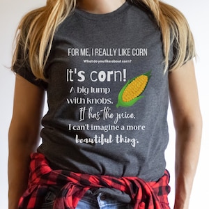 Può includere: Una maglietta grigia con una grafica bianca di una pannocchia di mais e il testo "Per me, mi piace davvero il mais. Cosa ti piace del mais? È mais! Un grosso grumo con pomelli. Ha il succo. Non riesco a immaginare qualcosa di più bello."
