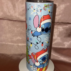 Puede incluir: Un vaso de acero inoxidable con un dise&ntilde;o festivo que presenta a Stitch de Lilo & Stitch. El vaso azul claro tiene copos de nieve, luces navide&ntilde;as y Stitch con un gorro de Pap&aacute; Noel. Se muestra sobre un pedestal blanco.