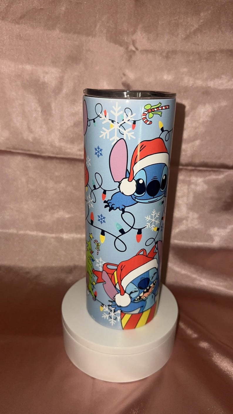 Puede incluir: Un vaso de acero inoxidable con un dise&ntilde;o festivo que presenta a Stitch de Lilo & Stitch. El vaso azul claro tiene copos de nieve, luces navide&ntilde;as y Stitch con un gorro de Pap&aacute; Noel. Se muestra sobre un pedestal blanco.