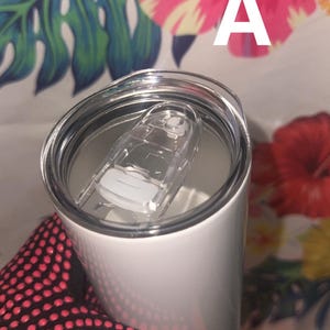 Puede incluir: Un vaso t&eacute;rmico blanco con tapa de pl&aacute;stico transparente y cierre deslizante. El vaso est&aacute; sostenido por una mano que lleva un guante negro con lunares rosas. El fondo presenta un estampado floral con la letra "A".