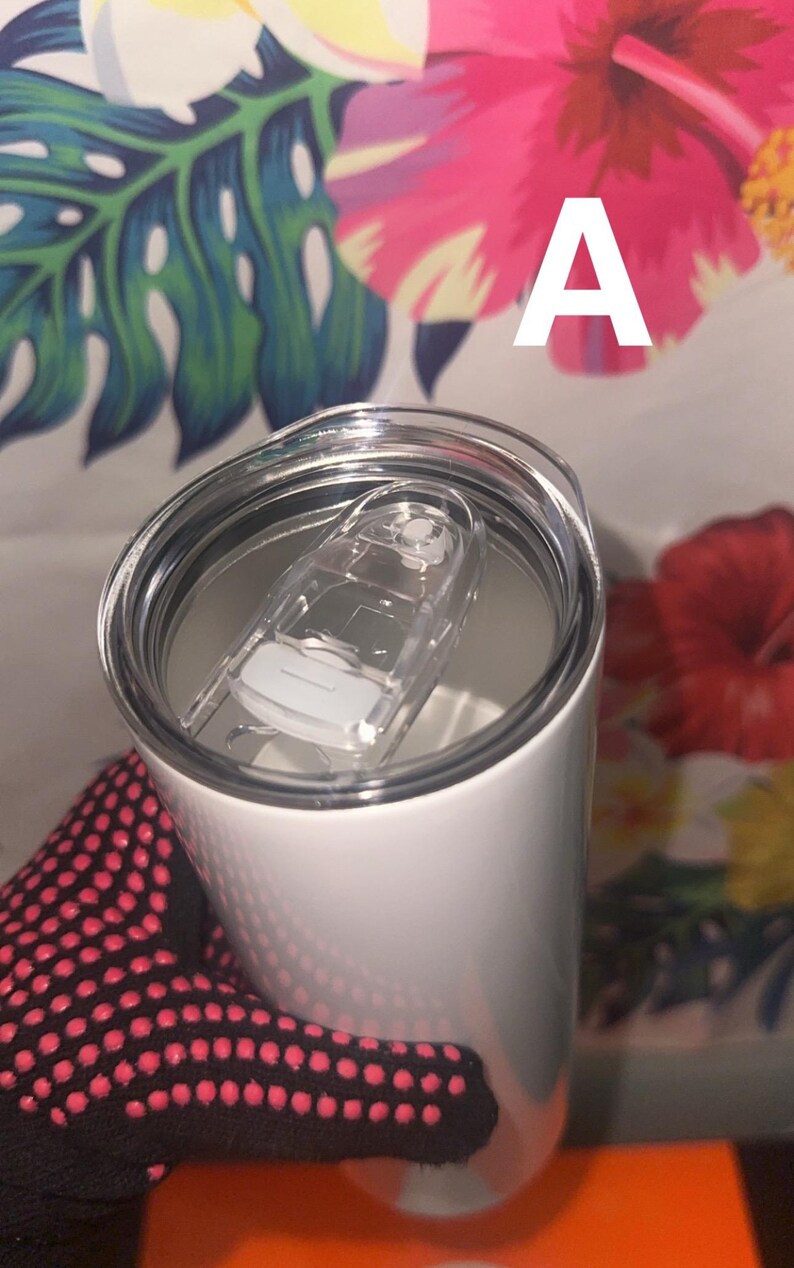 Puede incluir: Un vaso t&eacute;rmico blanco con tapa de pl&aacute;stico transparente y cierre deslizante. El vaso est&aacute; sostenido por una mano que lleva un guante negro con lunares rosas. El fondo presenta un estampado floral con la letra "A".